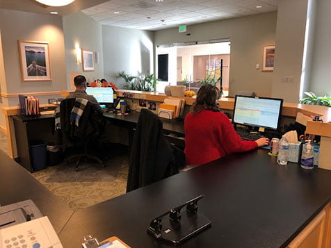 Office workers at desks with computers, in a brightly lit room with a reception counter in the foreground.