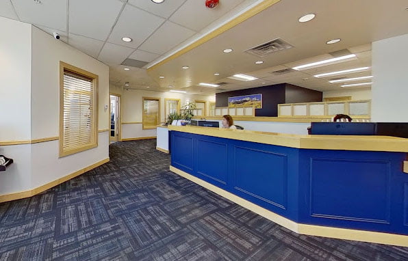 Interior of a modern office with blue reception counters, gray carpeted floor, and walls decorated with photos. two people are present, working at the counters.