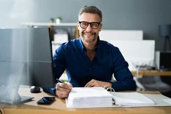 Smiling middle-aged man in glasses and a blue shirt, sitting at a desk in an office, writing notes on a stack of papers.