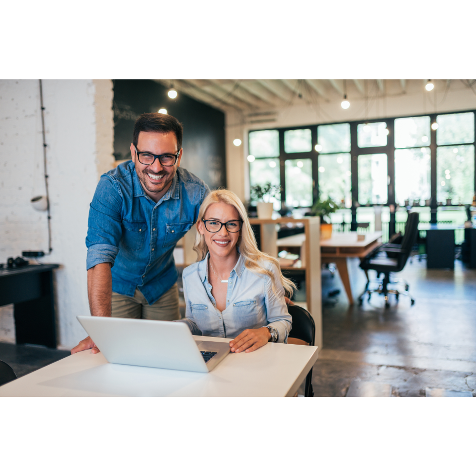 A man and a woman smiling at the camera, using a laptop in a modern office with large windows and brick walls.