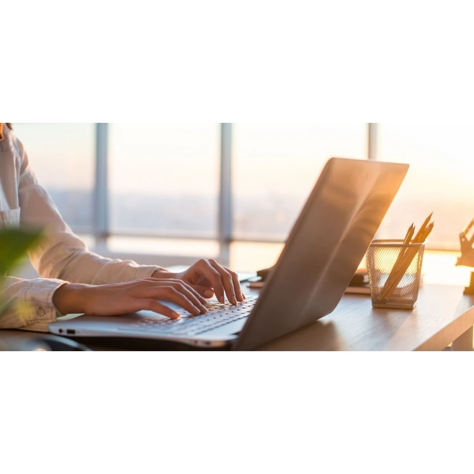 A person typing on a laptop at a desk with a sunset view outside the window, indicating a modern office environment.