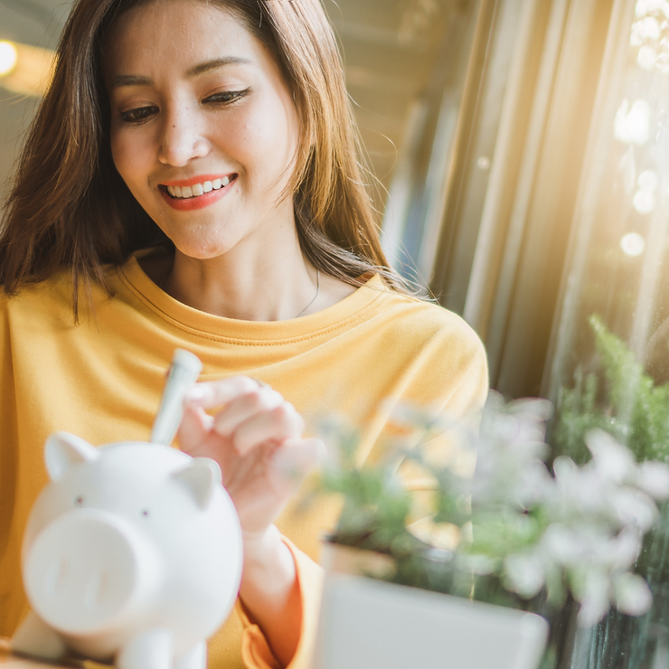 A smiling woman in a yellow sweater inserting a coin into a white piggy bank, standing beside indoor plants in a sunlit room.