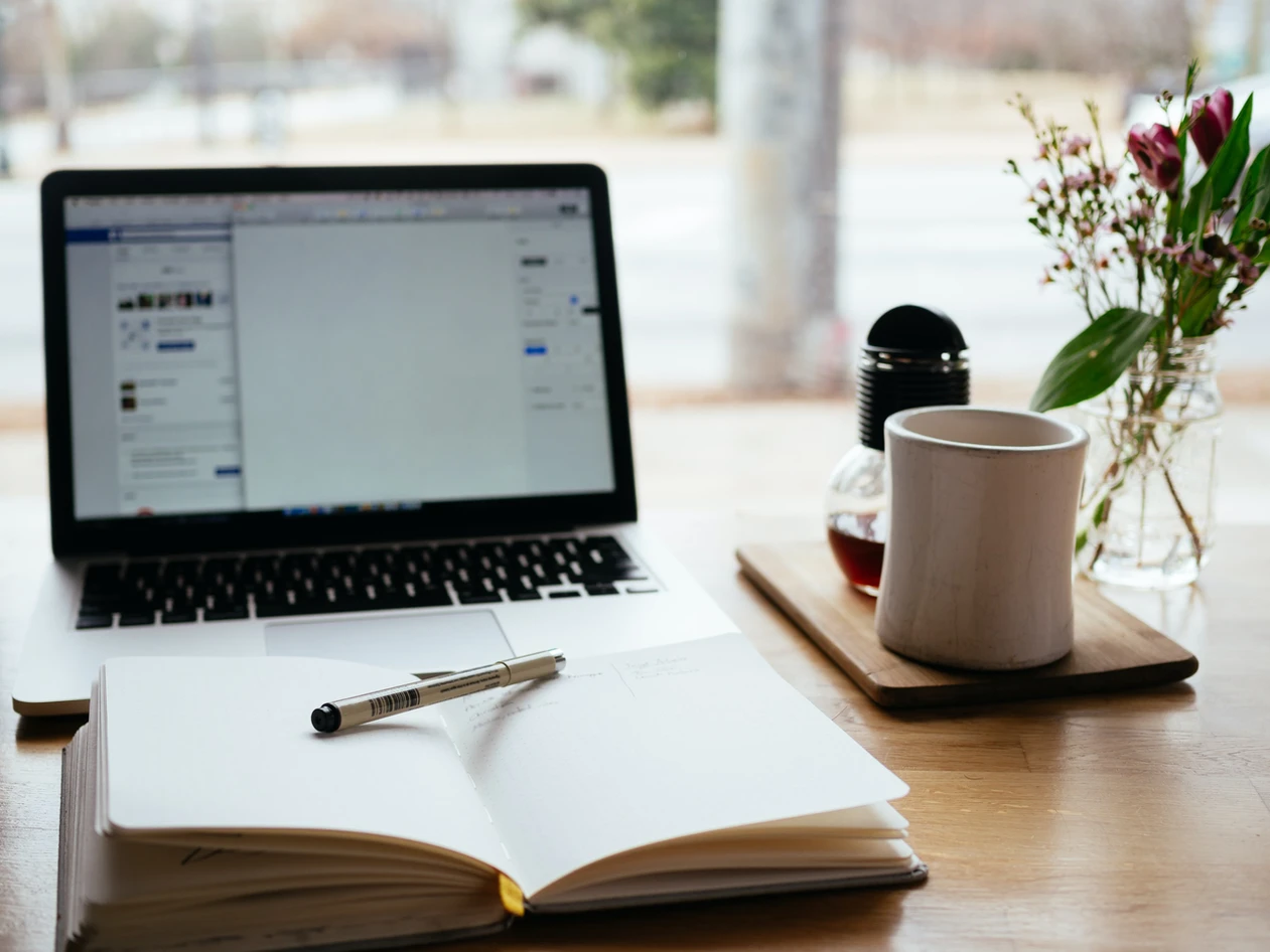 Laptop open on facebook with a blank notebook, pen, and a mug on a desk, suggesting a work or study environment.