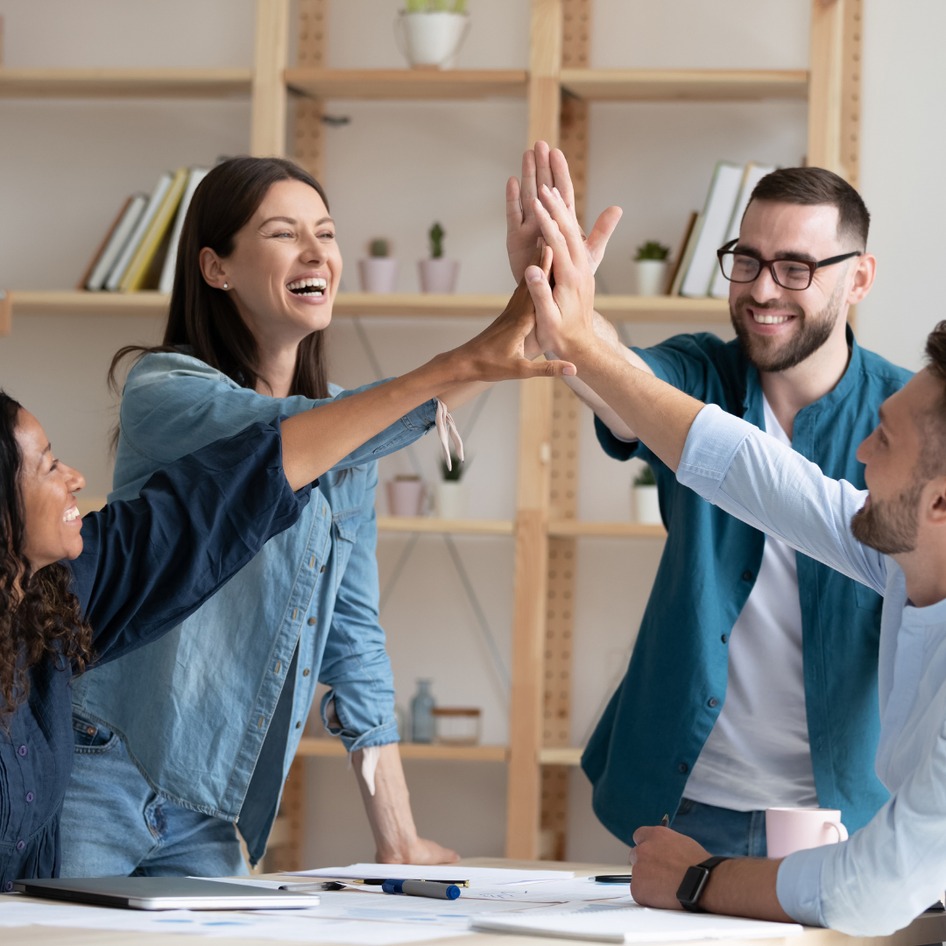 Four professionals giving each other a high-five in a meeting room, smiling and collaborating in a supportive team environment.