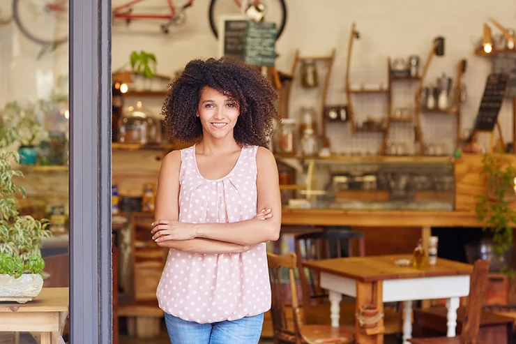 A confident woman with curly hair smiling while standing with crossed arms in the doorway of a cozy, well-lit café.