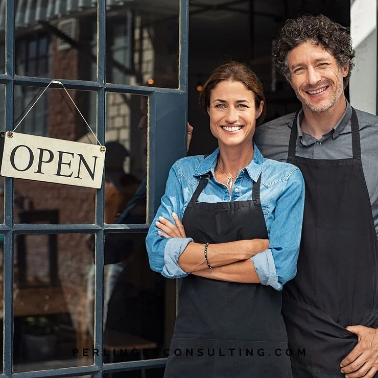 Two smiling small business owners stand in front of their shop, with an "open" sign hanging on the door.