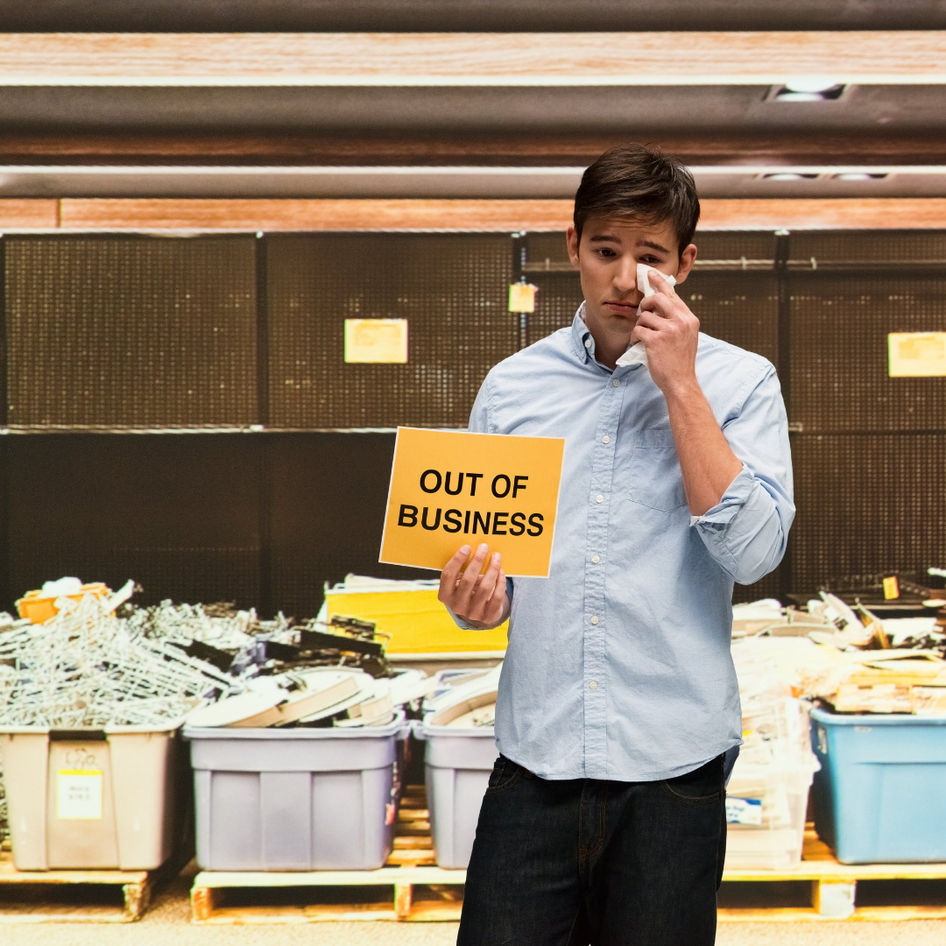 A young man talking on a cellphone holds a sign saying "out of business" in a cluttered, empty store.