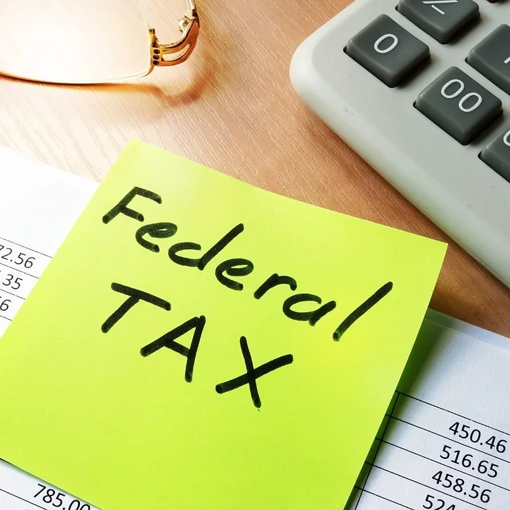 A green sticky note with "federal tax" handwritten on it, placed on a table next to a calculator and financial documents.