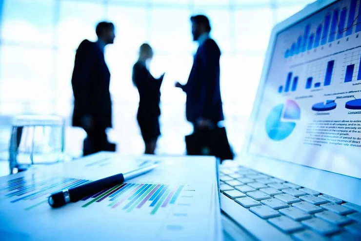 Silhouetted businesspeople discussing in an office, with focus on a foreground laptop displaying graphs and a financial report.