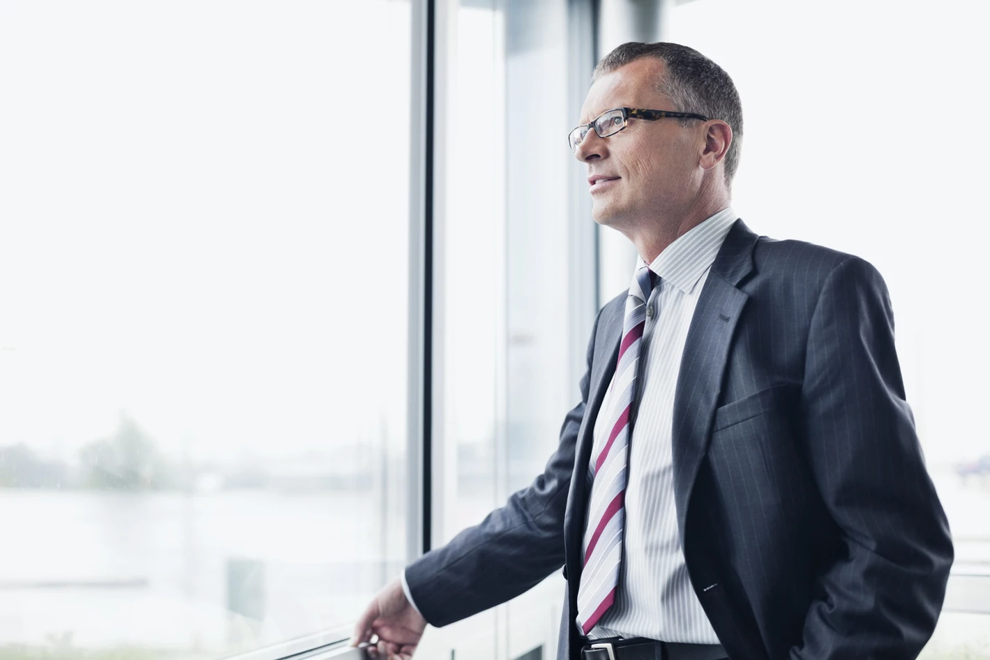 A middle-aged businessman in a suit and glasses looks thoughtfully out a window in a modern office.