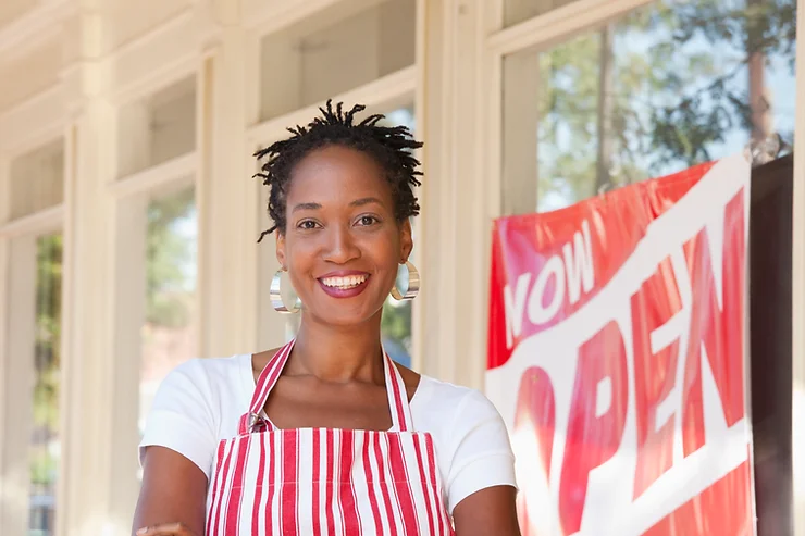 A smiling black woman wearing a red and white striped apron stands in front of a "now open" sign at a sunny storefront.