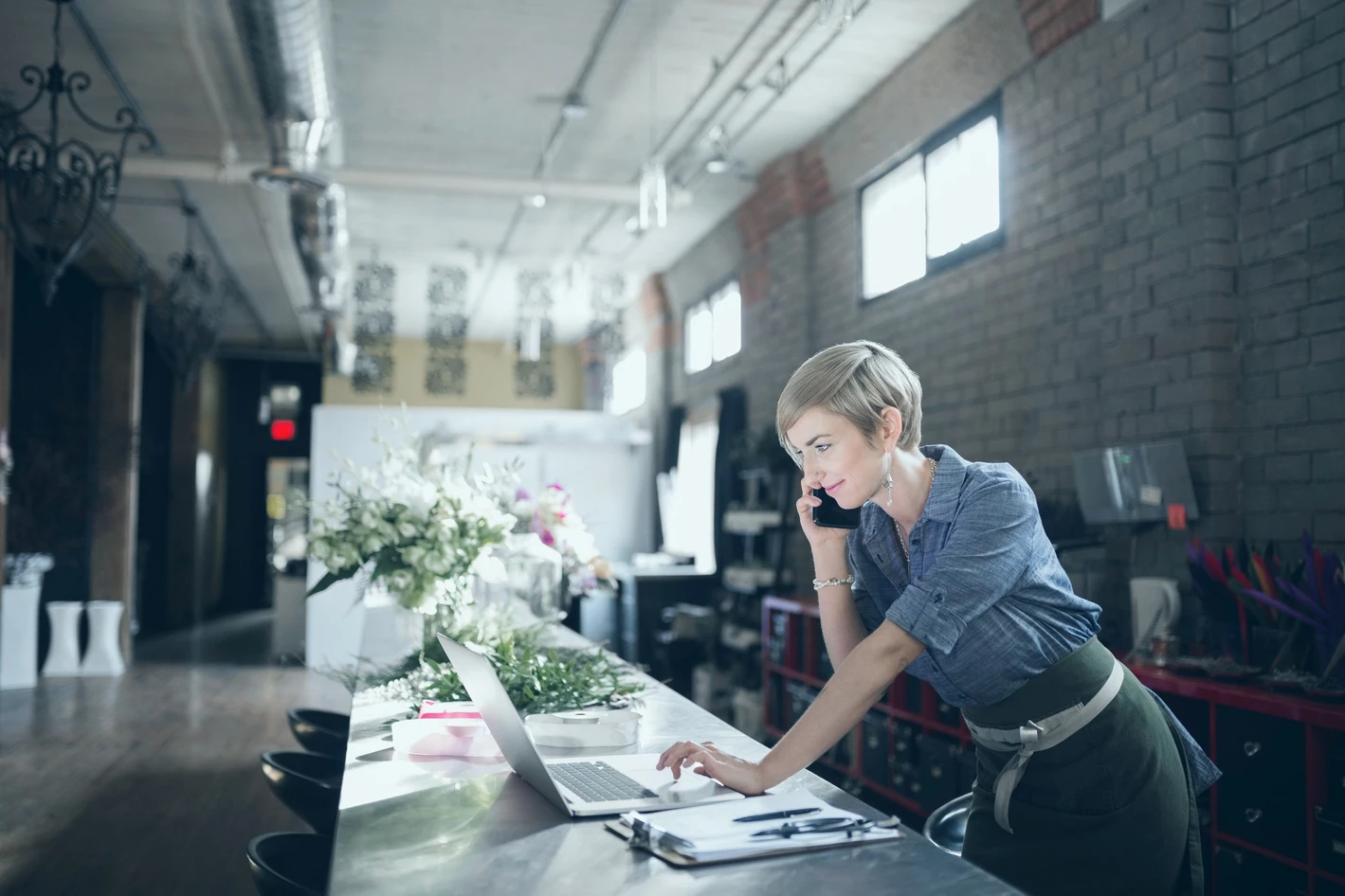 A woman in a denim shirt speaks on the phone while using a laptop in a modern loft-style office with exposed brick walls and large windows.