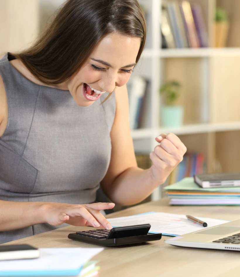 Woman in a gray dress excitedly raises a fist while looking at a calculator on a desk filled with papers and a laptop.