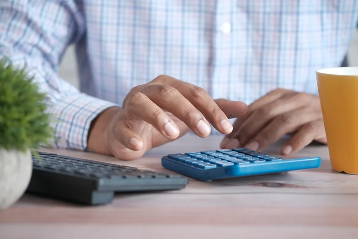 A person in a plaid shirt using a calculator on a desk with a keyboard, plant, and coffee cup nearby.