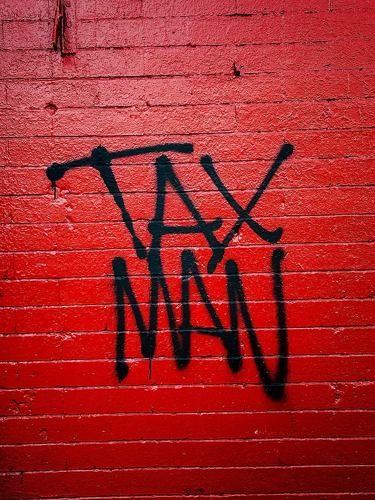 Graffiti spelling "tax" sprayed in black paint on a vibrant red brick wall.