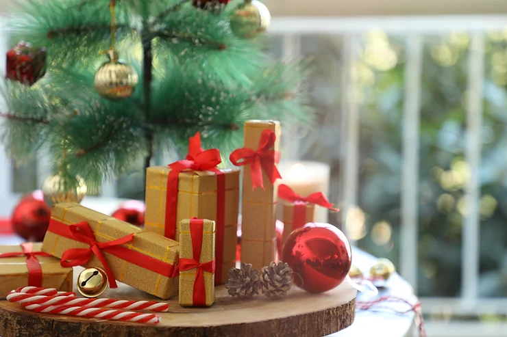 Decorative christmas setting with wrapped gifts, red and gold baubles, and candy canes placed on a wooden surface near a pine tree.