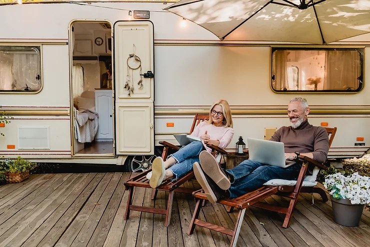 A mature couple relaxes in chairs outside a camper, the woman reading a book and the man using a laptop.