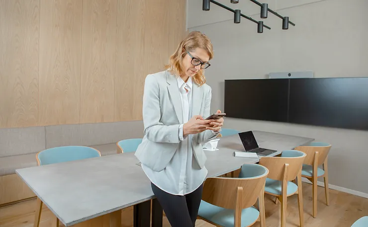 A woman in a light gray suit using a smartphone in a modern meeting room with a wooden table, blue chairs, and a laptop.