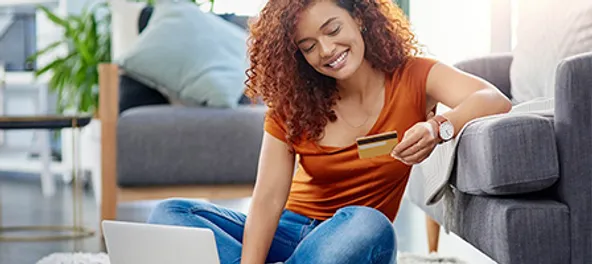 A smiling woman sitting on the floor uses a laptop and holds a credit card, likely shopping online. she's in a cozy living room.