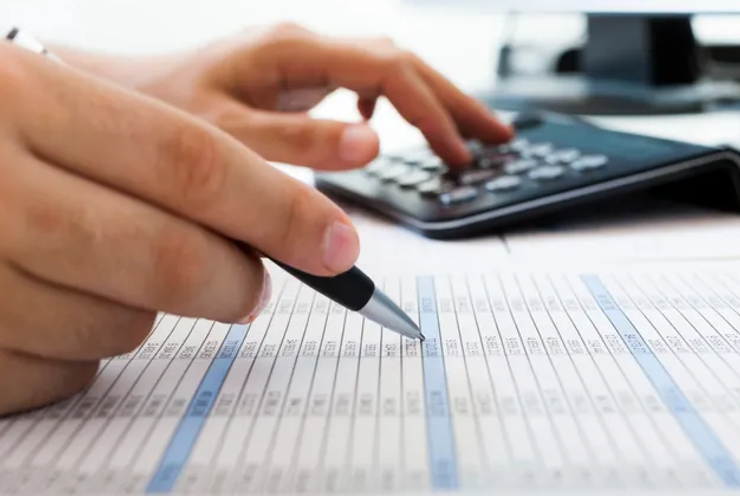 A person's hand holding a pen, pointing at a document with numbers, with a calculator nearby on a desk.