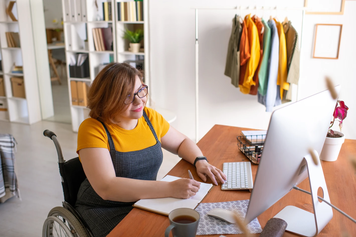 A woman in a wheelchair working at a desk with a computer, papers, and a coffee cup in a bright office setting.