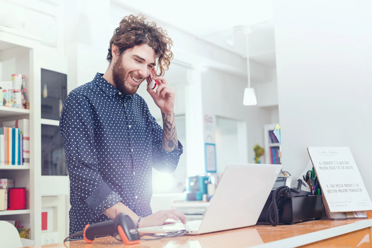 Man with curly hair smiling while talking on the phone and working on a laptop in a bright, cluttered office space.