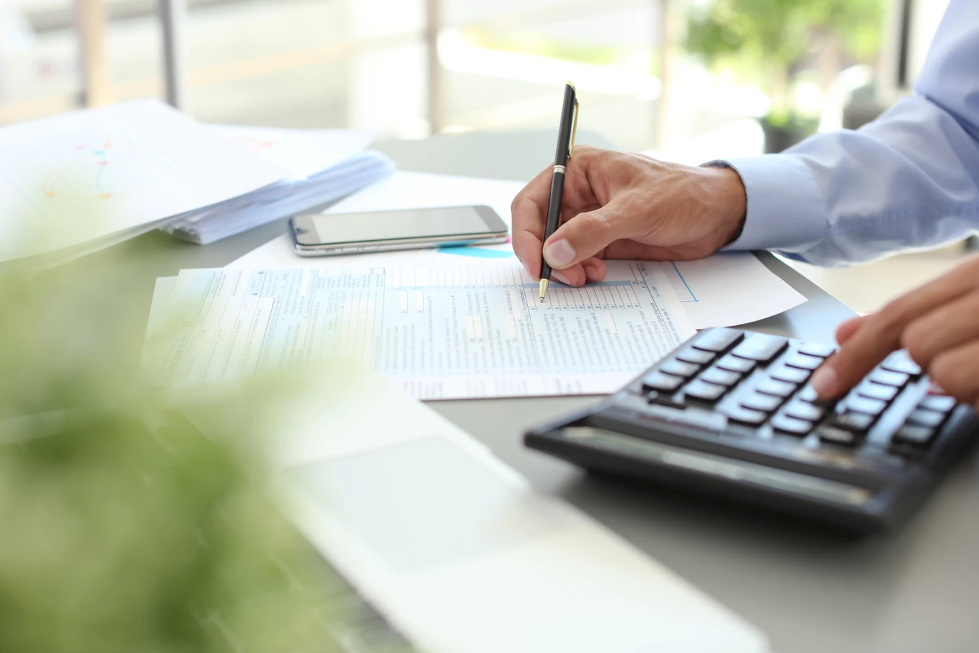 A person calculating expenses using a calculator and taking notes on a document at a desk, with a smartphone nearby.