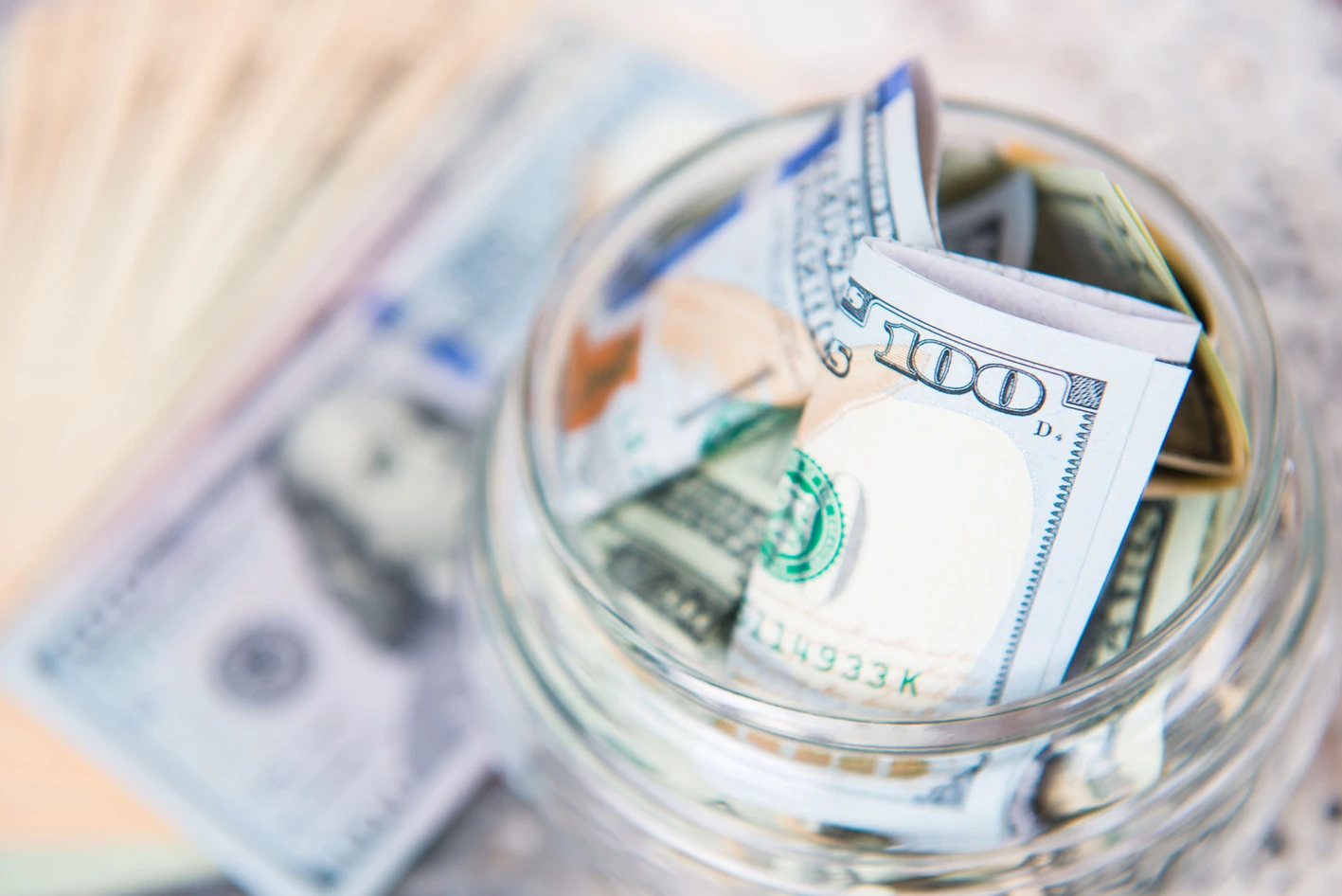 U.s. hundred-dollar bills crumpled and placed in a clear glass jar with a blurry background of additional banknotes.