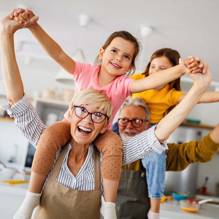 A joyful family in a kitchen with a grandmother in glasses and an apron, a grandfather, and two young girls all smiling brightly.