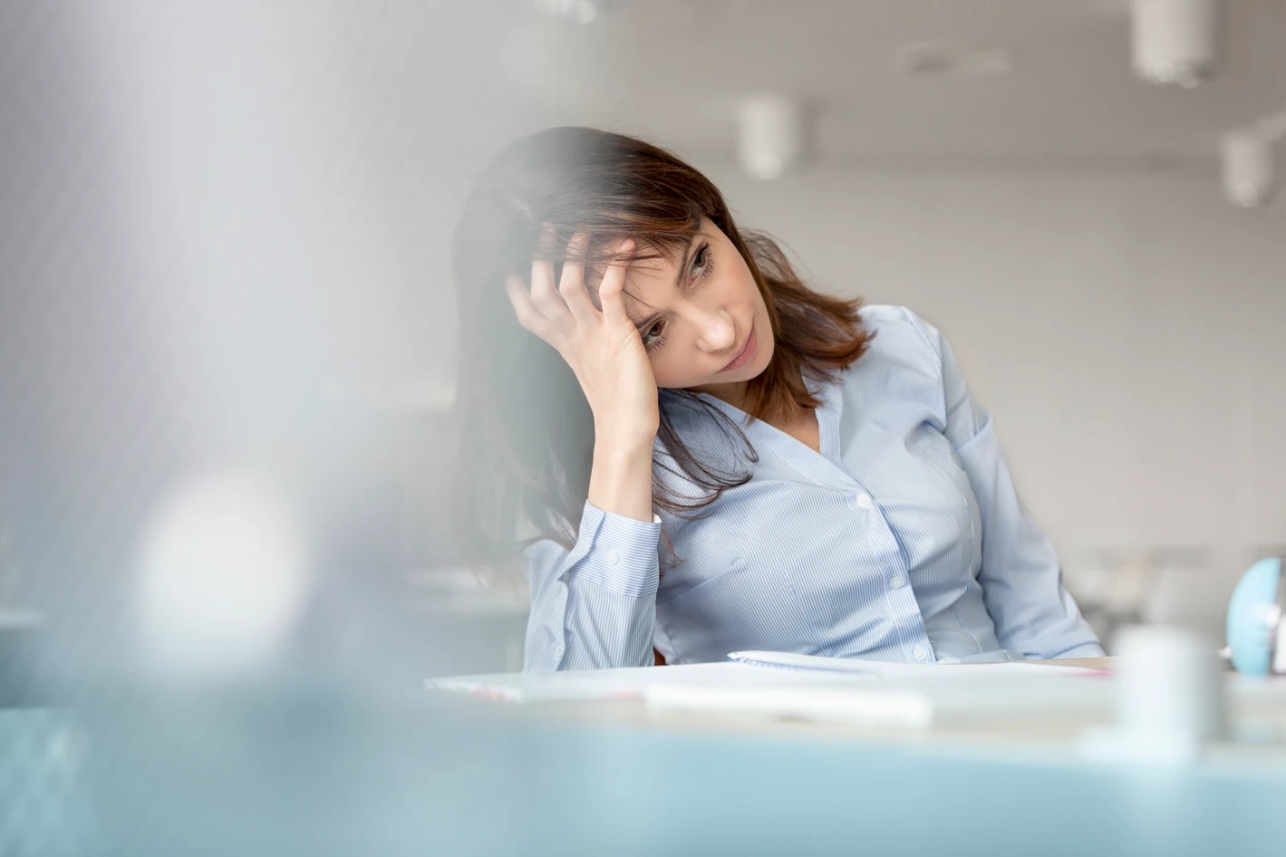 A woman with a contemplative expression sitting at a desk, resting her head on her hand, with papers in front of her