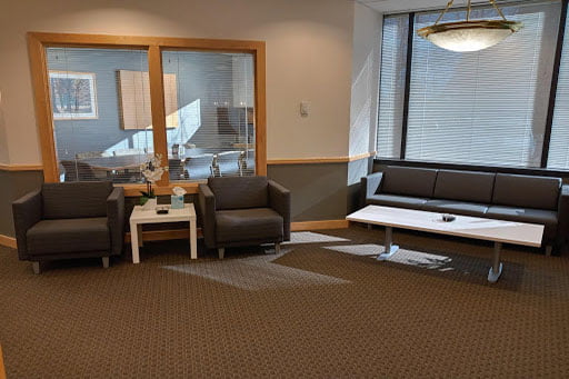 Modern office lounge with a gray sofa, two armchairs, a coffee table, and a glass-partitioned meeting room in the background.