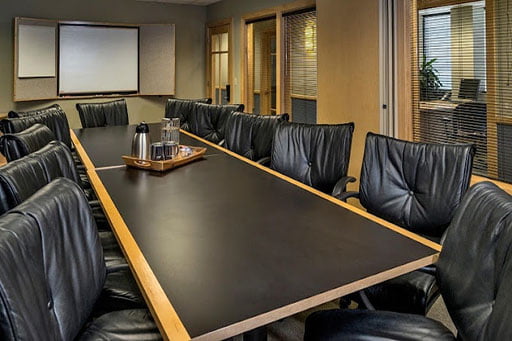 Empty conference room with a long table surrounded by black chairs and a whiteboard on the back wall.