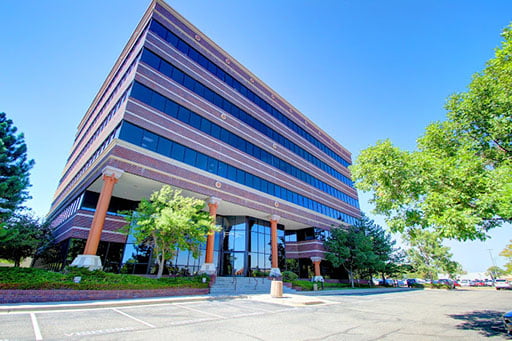 Low-angle view of a modern multi-story office building with large pillars and a glass facade under a clear blue sky.