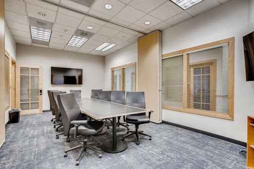 Modern office meeting room with a long table, gray chairs, wooden floor, and large windows with blinds.