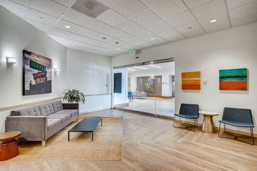 Modern office lobby with a gray sofa, two accent chairs, a coffee table, and colorful wall art, leading to a glass door entryway.